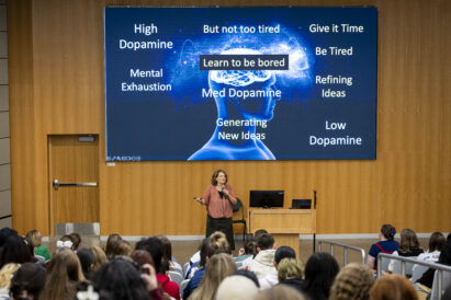 A speaker addresses an audience in a lecture hall, standing before a large screen displaying a diagram about dopamine levels and stages of creative thinking.